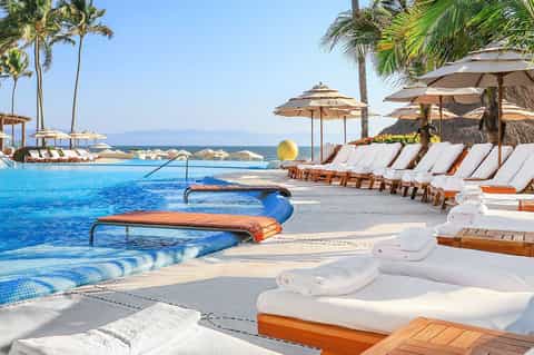 Luxury resort pool with ocean view, white loungers, umbrellas, palm trees, and distant mountains