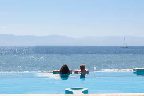Infinity pool overlooking ocean with two swimmers, sailboat visible on water