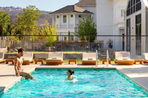 Contemporary pool area with white loungers, glass railing, modern white mansion, and hillside views