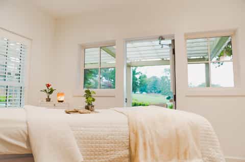 Bright bedroom with white linens, large windows overlooking green landscape, and minimalist décor