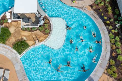 Overhead view of curved resort pool with swimmers, white umbrellas, and landscaped grounds