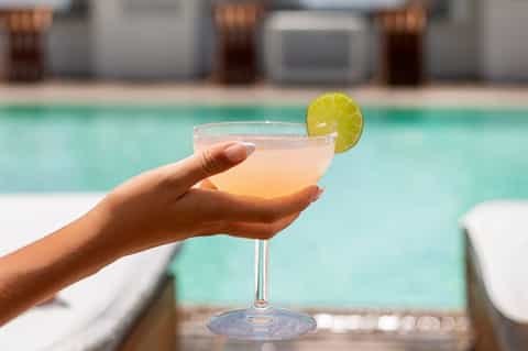 Hand holding tropical cocktail with lime wheel by turquoise pool and loungers