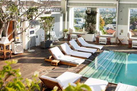 Rooftop pool deck with wooden lounge chairs, white cushions, and panoramic city skyline views through large windows
