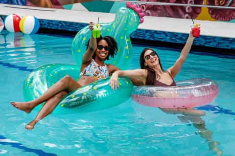 Two women on inflatable pool floats with drinks at resort pool party