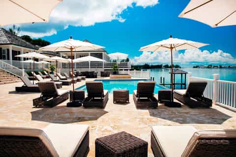 Beachfront pool with brown lounge chairs, white umbrellas, and turquoise water with dock views.