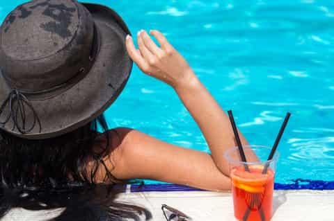 Person relaxing by hotel pool wearing hat with colorful drink, turquoise water visible