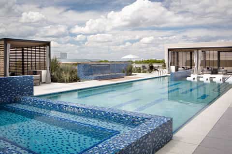 Rooftop pool with blue tile, cabanas with shutters, loungers, and city skyline views