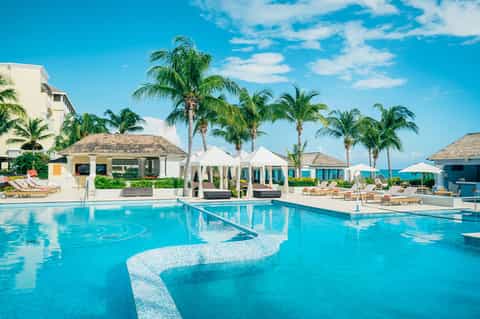 Tropical resort pool with turquoise water, palm trees, white cabanas, lounge chairs, and ocean backdrop