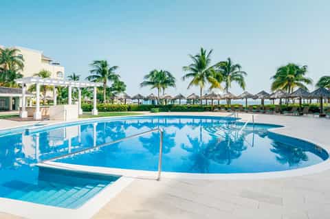 Curved resort pool with white pergola, palm trees, and beach umbrellas in background