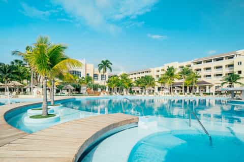 Large resort swimming pool with palm trees, multi-level pool areas, and yellow building accommodations