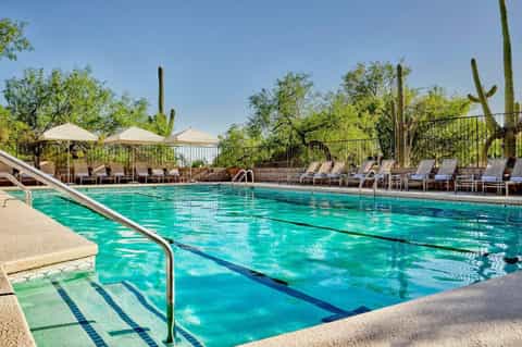 Clear lap pool with lane markers, tan umbrellas, lounge chairs, and desert saguaro cacti in background