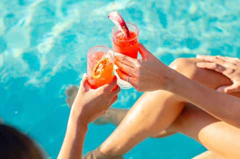 Two people toasting with colorful cocktails while relaxing by turquoise swimming pool