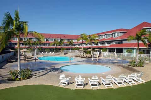 Resort courtyard with large rectangular pool, hot tub, white loungers, red-roofed building, and palm trees