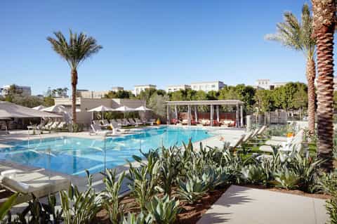 Resort swimming pool with palm trees, white loungers, pergolas, and clear blue sky
