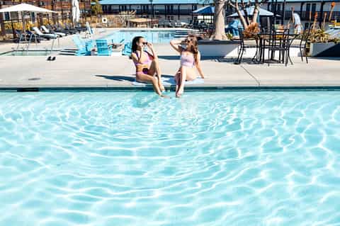 Resort pool area with lounge chairs, blue umbrellas, decorative trees, and guests enjoying the water