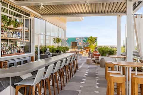 Outdoor rooftop bar with modern pergola, bar seating, lounge furniture, ocean views, and tropical plants