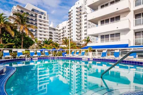 Resort pool with turquoise water, blue lounge chairs, palm trees, and multi-story beachfront hotel buildings