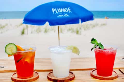 Three colorful cocktails with fresh garnishes on a wooden deck under a blue Plunge umbrella, beach and ocean in background