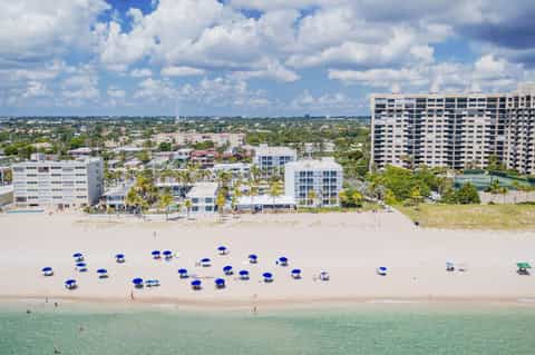 Aerial view of beachfront resort with blue umbrellas on sand, modern buildings, and turquoise ocean waters