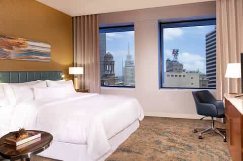 Luxury hotel bedroom with king bed, city skyline views, warm gold walls, and contemporary furnishings