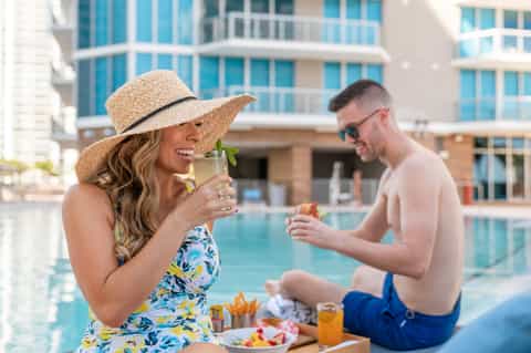 Woman in hat holding cocktail by pool with man, resort buildings in background