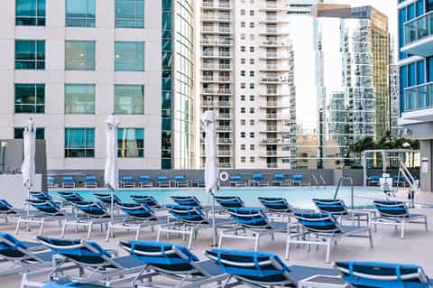 Urban rooftop pool with blue lounge chairs, white umbrellas, and high-rise residential buildings