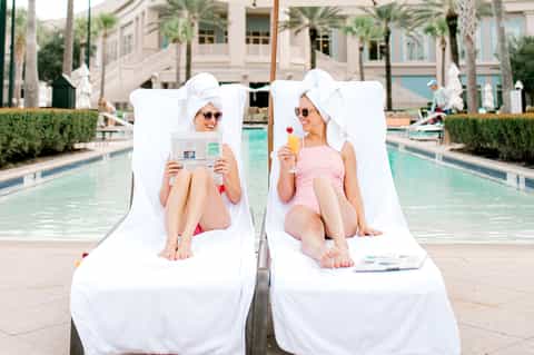 Two women lounging poolside in white chairs with beverages and newspaper