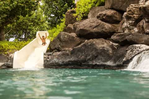 Child on white water slide at tropical resort pool surrounded by natural rock formations and lush greenery.