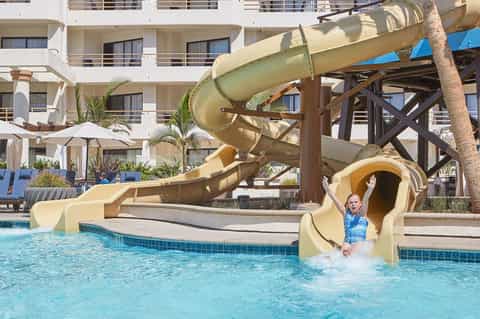 Child exiting yellow water slide into turquoise pool at resort with white hotel building