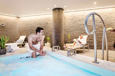 Indoor spa pool with stone column, man in water, lounge chairs, and rolled towels in modern wellness center
