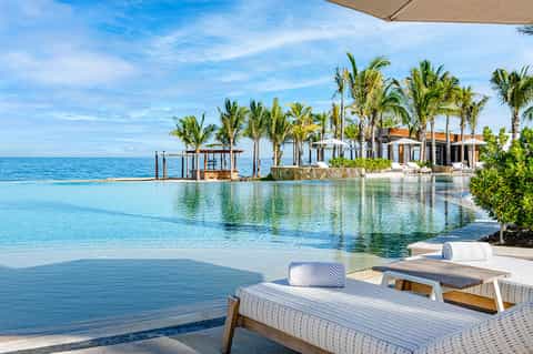 Luxury beachfront infinity pool with palm trees, turquoise water, white lounge chairs, and ocean horizon view