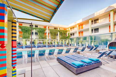 Colorful hotel courtyard with striped awning, lounge chairs, and modern residential buildings surrounding a sparkling pool.