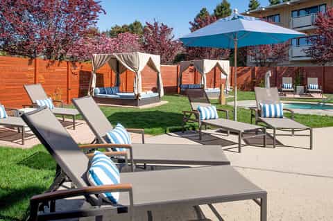 Resort pool area with white loungers, blue umbrellas, and enclosed cabanas surrounded by landscaping