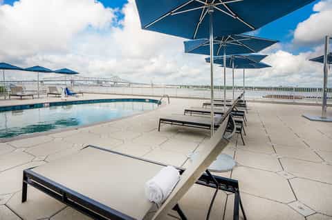 Rooftop pool with blue umbrellas, loungers, river view, and architectural bridge in background
