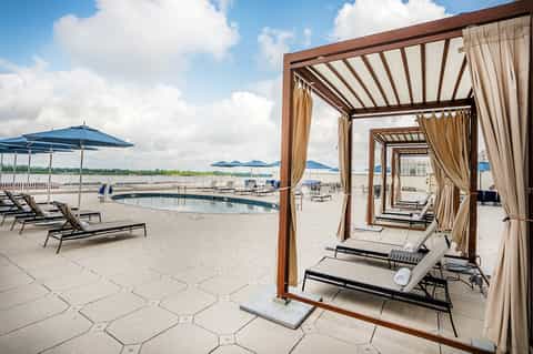 Waterfront pool deck with brown pergolas, tan curtains, blue umbrellas, and water view