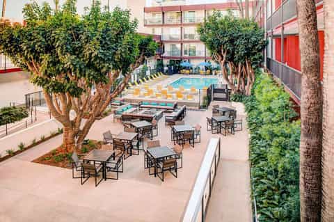 Courtyard pool area surrounded by tall trees, lounge seating, and colorful umbrellas in urban resort setting