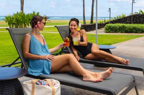 Two women relaxing on lounge chairs with tropical ocean and palm tree views, enjoying beverages