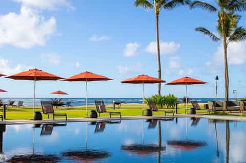 Resort pool area with orange umbrellas, lounge chairs, palm trees, manicured lawn, and ocean horizon