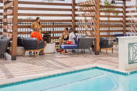 Resort guests relaxing in lounge chairs under striped wooden pergola by infinity pool and beach