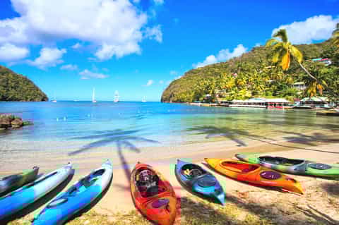 Tropical beach cove with colorful kayaks on sand, sailboats on turquoise water, palm trees and lush hills