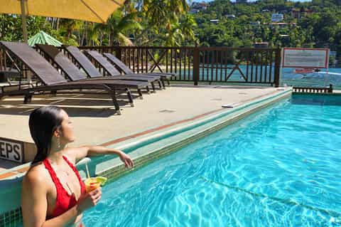 Woman in red swimsuit at pool edge overlooking turquoise water with wooden deck, lounge chairs, and railing