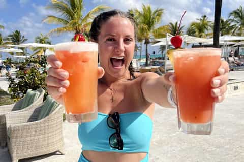 Woman holding two colorful tropical drinks at a beach bar with palm trees in background