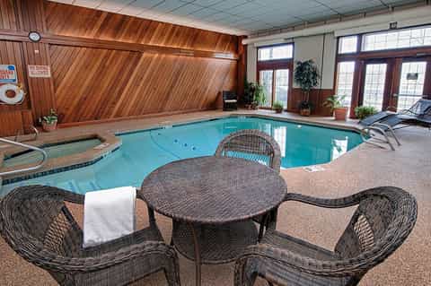 Indoor pool facility with wooden vaulted ceiling, turquoise water, lounge seating, and potted plants.