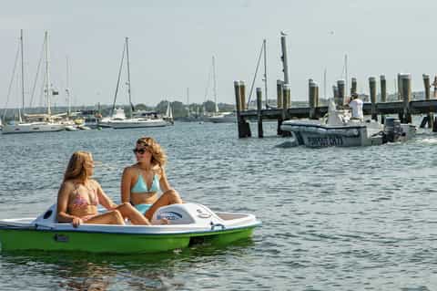 Two women on green motorboat with sailboats and dock pilings visible in calm harbor
