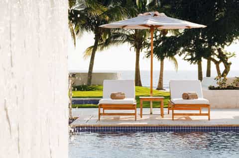 Poolside loungers with white cushions under wooden umbrella overlooking ocean and palm trees