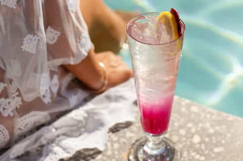 Tropical cocktail with pink and clear layers garnished with lemon and cherry by pool