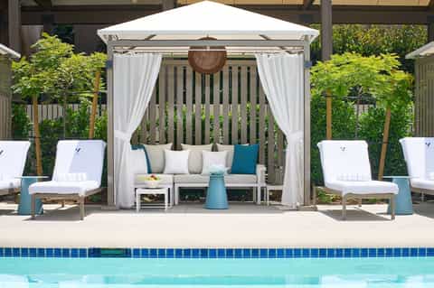 Luxury poolside cabana with white canopy, cream sofa, blue accent chairs, and tropical landscaping