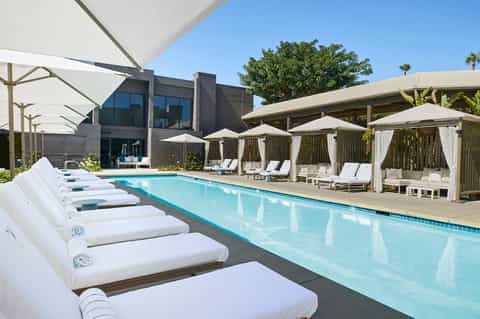 Resort pool lined with white lounge beds, cabanas with white curtains, and palm trees in bright sunshine