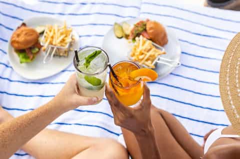 Hands holding refreshing beverages with burgers and fries on striped beach towel