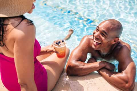 Couple in pool enjoying drinks, wearing pink and dark swimwear in clear blue water
